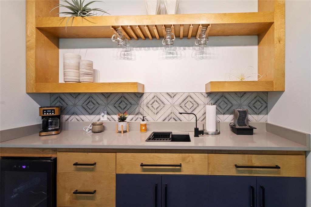 Built-in wet bar featuring custom two-tone cabinetry, solid-surface countertops, decorative tile backsplash, floating wooden shelves with integrated stemware rack, and a dedicated beverage refrigerator