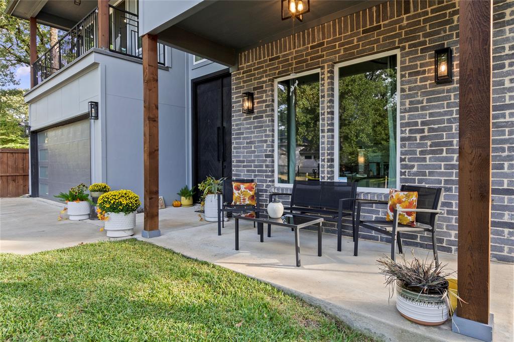 Covered patio featuring a grey brick facade, exposed wood-finish posts, double front doors, large windows, and wall-mounted exterior lighting