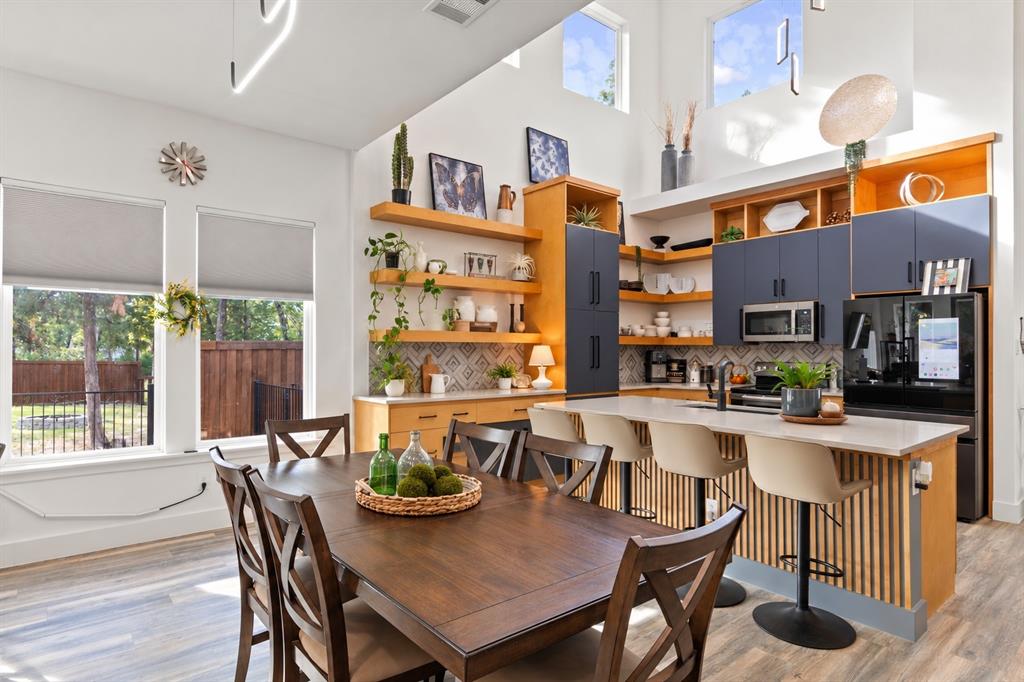 High-ceiling kitchen featuring a dual-toned cabinetry finish, wood-finish flooring, floating wood shelving, a central island with a slatted wood base, and geometric tile backsplash High-ceiling kitchen featuring a dual-toned cabinetry finish, wood-finish flooring, floating wood shelving, a central island with a slatted wood base, and geometric tile backsplash