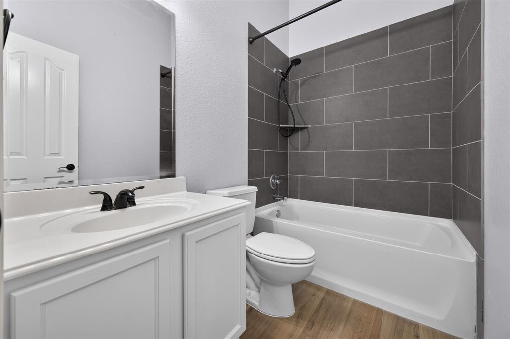 Bathroom featuring a white vanity cabinet with a solid surface countertop, matte black faucet hardware, wood-finish flooring, and a shower-tub combination with horizontal charcoal-toned tile surround