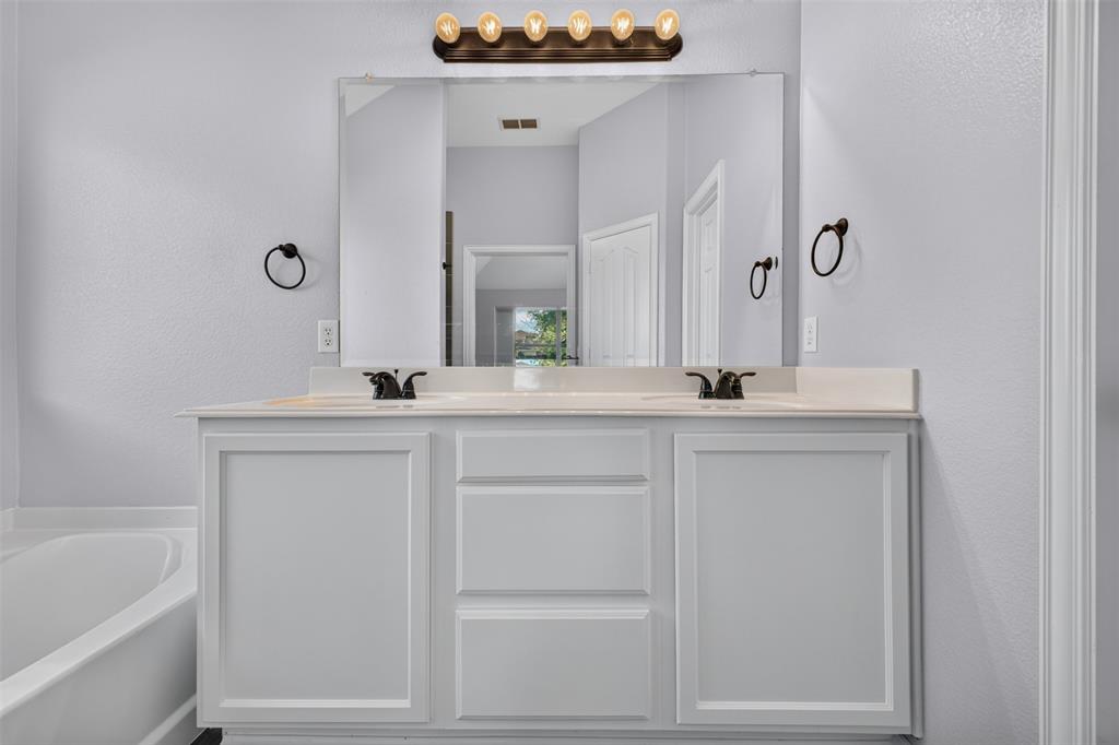 White dual-sink vanity featuring shaker-style cabinetry, solid-surface countertop, bronze-finish hardware, and a large frameless mirror with five-light vanity fixture