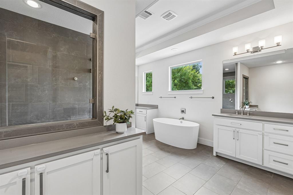 Freestanding soaking tub, double-sink vanity with quartz countertops, large-format tile flooring, and walk-in shower with dark tile surround