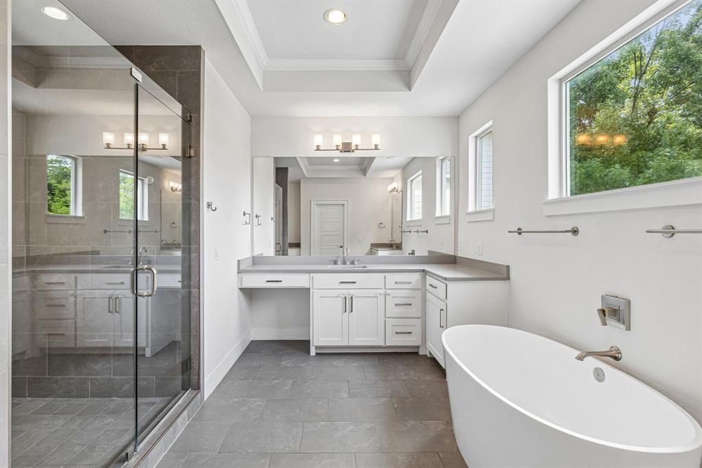 Freestanding soaking tub, frameless glass-enclosed shower with stone-finish surround, white cabinetry, solid-surface vanity countertop, and recessed tray ceiling