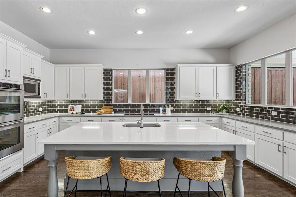Kitchen featuring a central island with a solid-surface countertop and undermount sink, dark brick-style tile backsplash, white shaker-style cabinetry, stainless steel double wall ovens, and wood-finish flooring