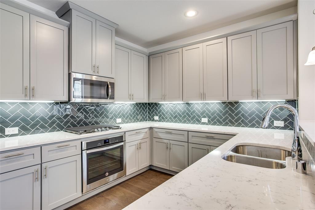 Kitchen featuring gray shaker-style cabinetry, white quartz countertops, a herringbone-patterned tile backsplash, stainless steel built-in appliances, and a dual-basin undermount sink Kitchen featuring gray shaker-style cabinetry, white quartz countertops, a herringbone-patterned tile backsplash, stainless steel built-in appliances, and a dual-basin undermount sink