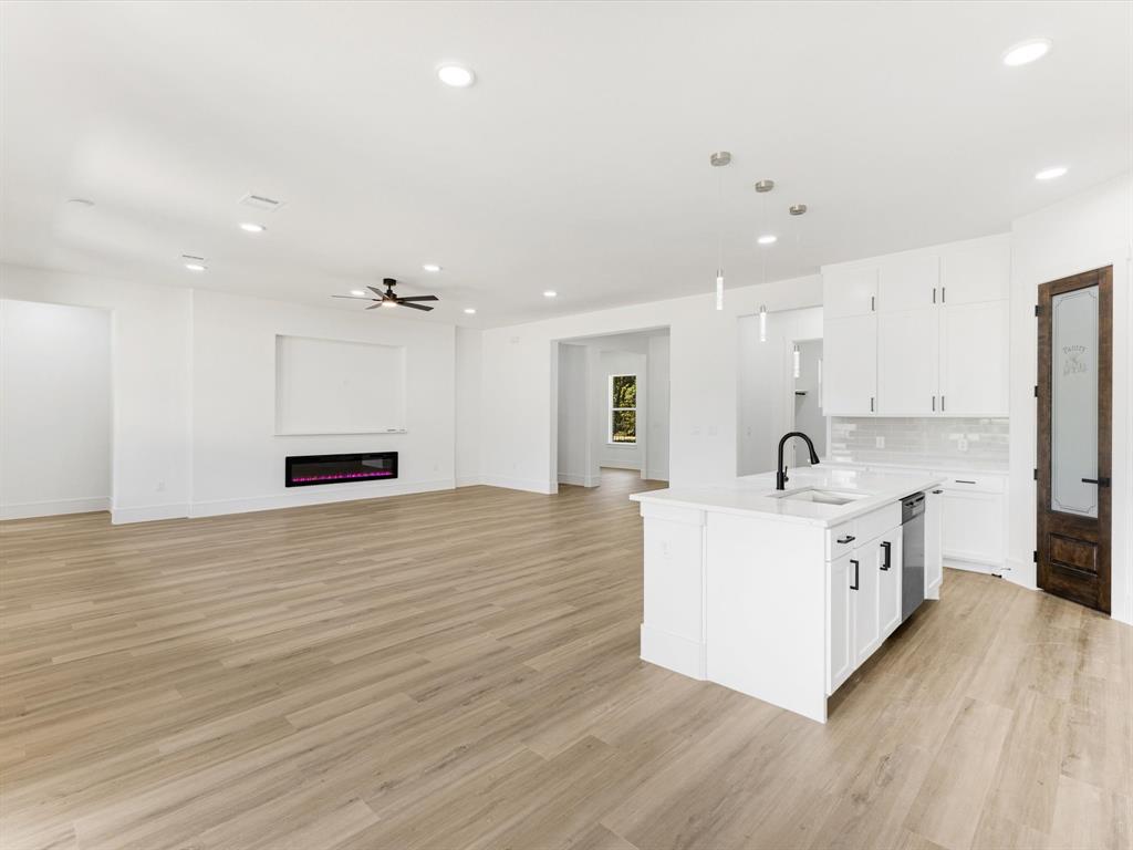 Open-concept interior featuring light wood-finish flooring, white cabinetry with matte black hardware, a central kitchen island with a matte black faucet, a built-in wall-mounted electric fireplace, and recessed lighting