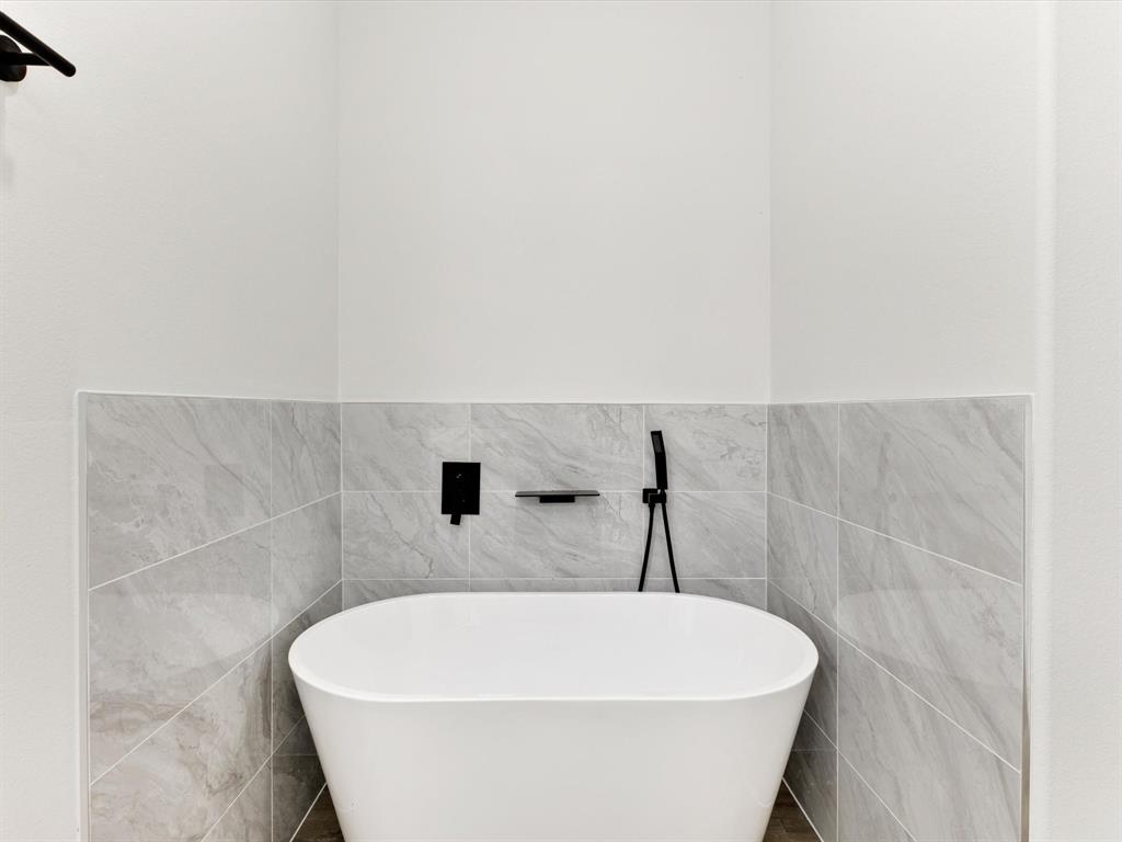 Freestanding white soaking tub with matte black wall-mounted faucet, handheld shower wand, built-in soap niche, and grey stone-finish tile surround