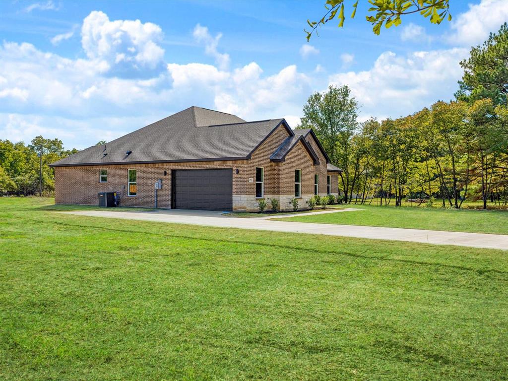 Brick and stone exterior featuring an attached multi-car garage, asphalt shingle roofing, and an expansive lawn on a wooded lot