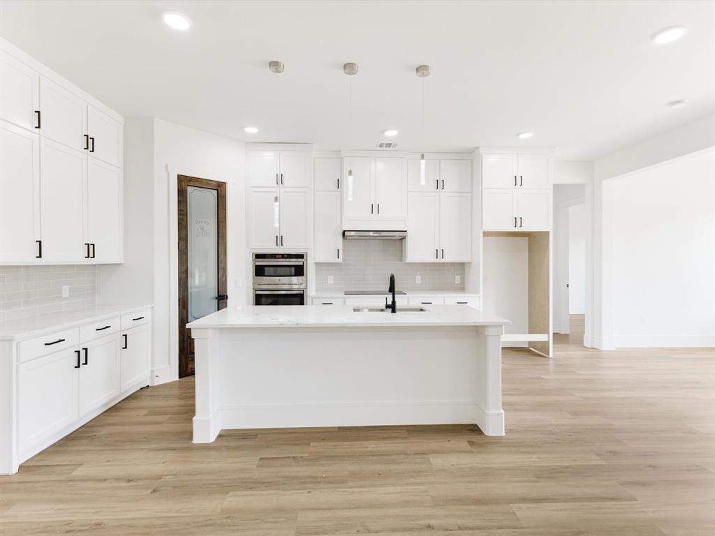 Kitchen featuring shaker-style cabinetry, a central island with a stone-finish countertop, stainless steel double wall ovens, wood-finish flooring, and a glass-paneled pantry door