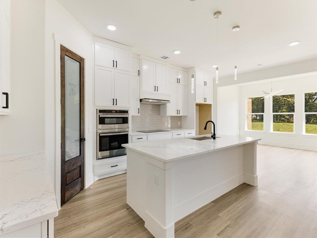 Kitchen featuring white shaker-style cabinetry, quartz countertops, a central island with an undermount sink, built-in double wall ovens, and a stained-wood pantry door with glass inlay