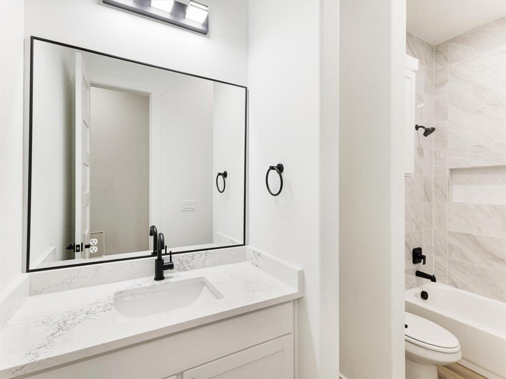 Bathroom vanity featuring a stone-composite countertop with an integrated rectangular sink, black-finish faucet, framed wall-mounted mirror, and tile-surround tub area with a built-in recessed niche