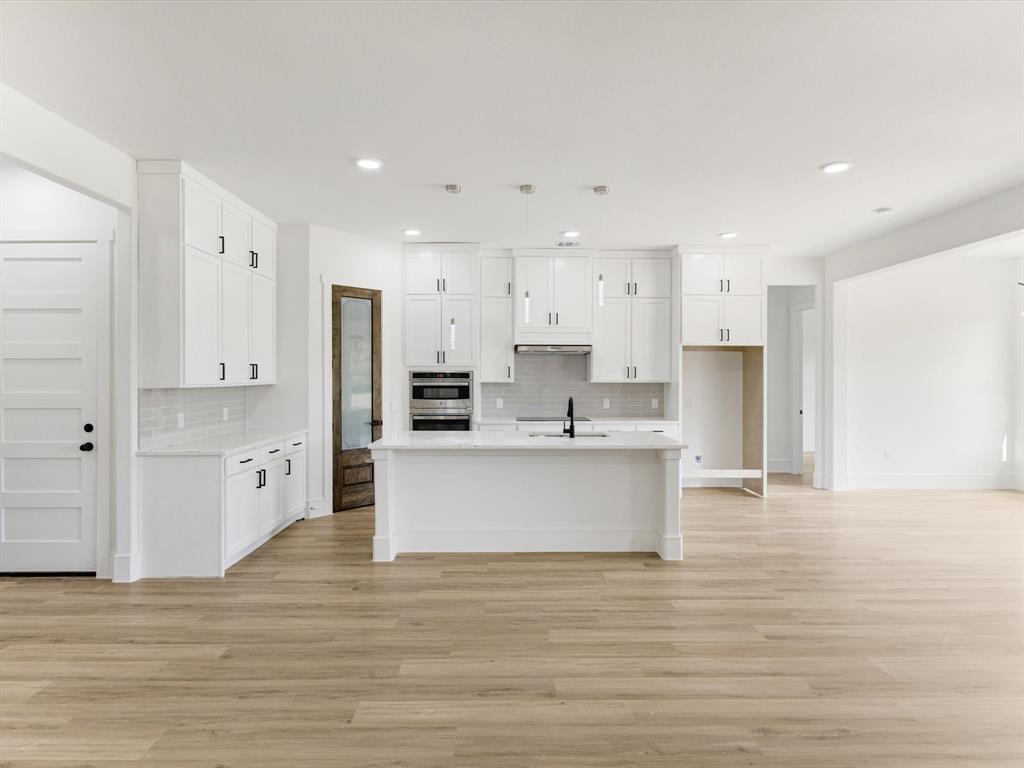 Kitchen featuring white ceiling-height cabinetry, a central island with a stone-finish countertop, wood-finish flooring, built-in stainless steel wall oven, and a pantry with a wood-toned door