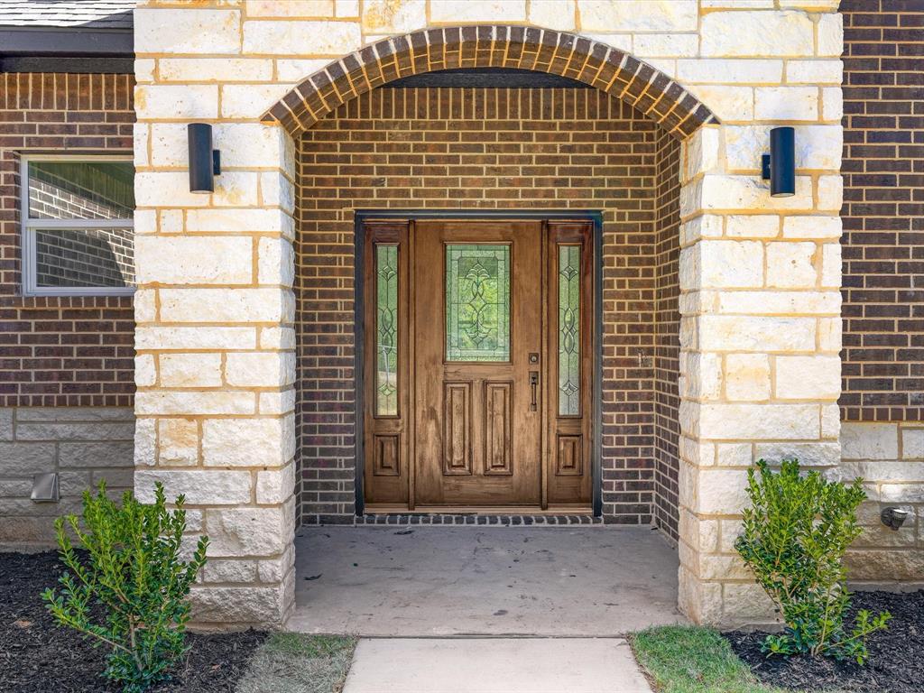 Entryway featuring a arched stone portico, brick facade, custom wood door with decorative glass paneling, dual sidelights, and exterior wall-mounted lighting