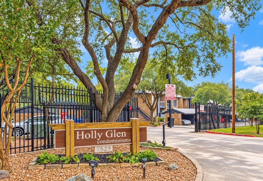Gated community entrance featuring metal perimeter fencing, a stone-bordered garden bed, mature tree canopy, and covered parking structures