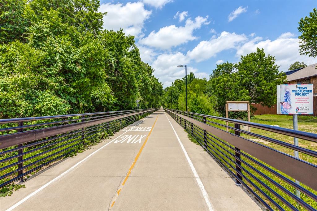 Paved multi-use trail featuring metal railings, overhead lighting, and mature perimeter trees