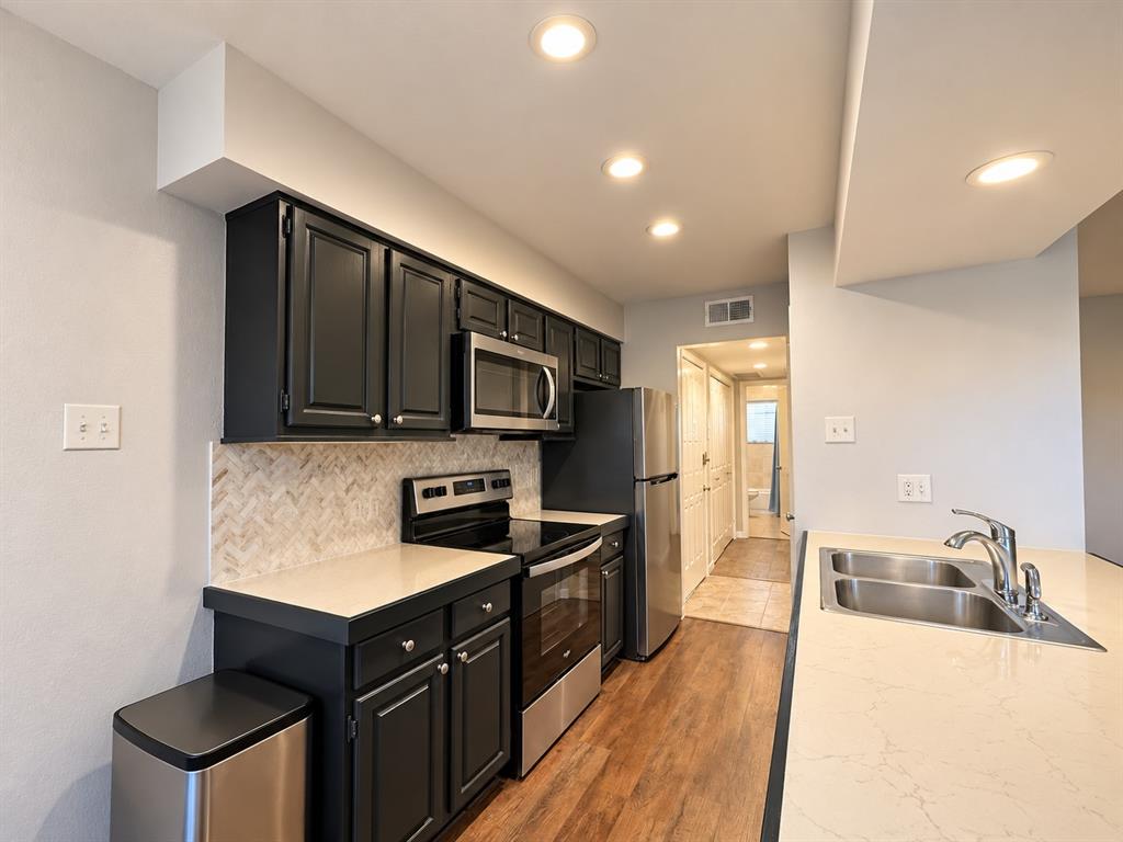 Kitchen featuring dark cabinetry, stone-finish countertops, stainless steel appliances, herringbone-patterned tile backsplash, and wood-finish flooring