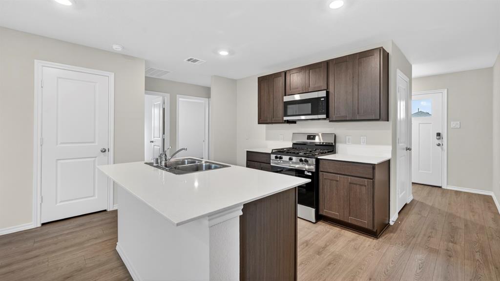 Kitchen featuring an island with a double basin sink, white countertops, dark wood-finish cabinetry, stainless steel microwave, and gas range Kitchen featuring an island with a double basin sink, white countertops, dark wood-finish cabinetry, stainless steel microwave, and gas range