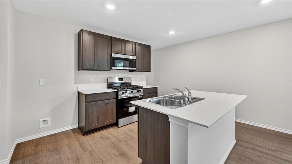 Kitchen featuring wood-finish flooring, dark wood-finish cabinetry, white countertops, stainless steel appliances, and a kitchen island with an integrated double sink Kitchen featuring wood-finish flooring, dark wood-finish cabinetry, white countertops, stainless steel appliances, and a kitchen island with an integrated double sink