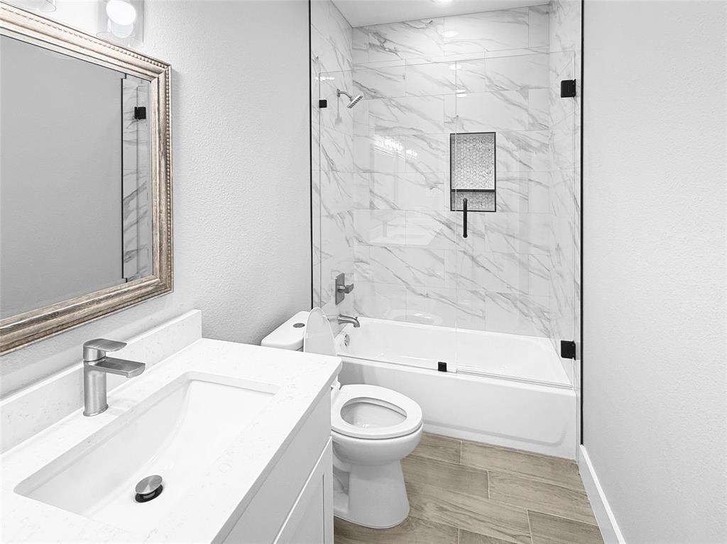 Bathroom featuring a vanity with an integrated rectangular sink and stone-finish countertop, wood-finish flooring, and a tub-shower combination with floor-to-ceiling tile surround, glass enclosure, and built-in recessed shelving