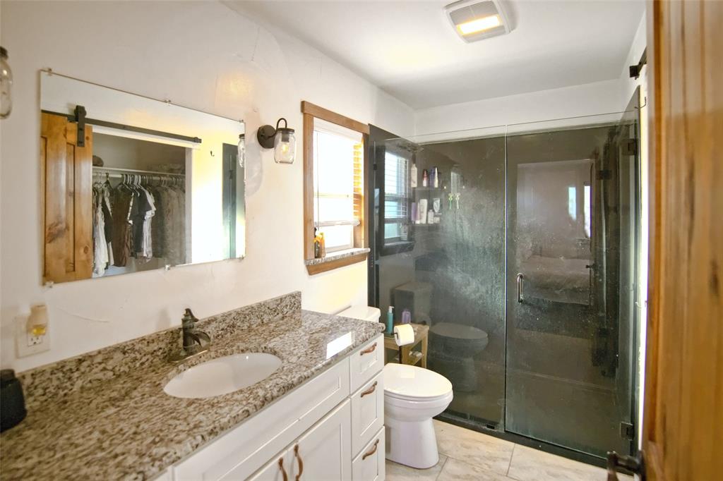 Stone-topped vanity featuring white cabinetry, glass-enclosed shower with dark metal hardware, wall-mounted light fixtures, and wood-finish trim