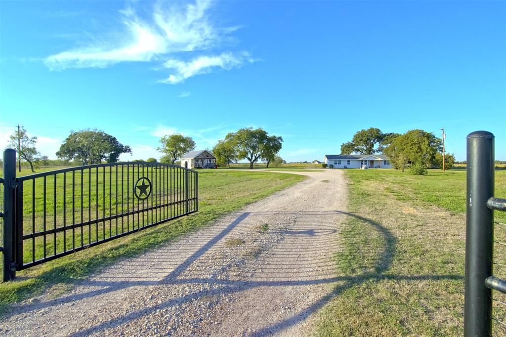 Gated entry featuring a decorative star motif and wrought-iron fencing, gravel driveway, and expansive acreage with mature trees Gated entry featuring a decorative star motif and wrought-iron fencing, gravel driveway, and expansive acreage with mature trees