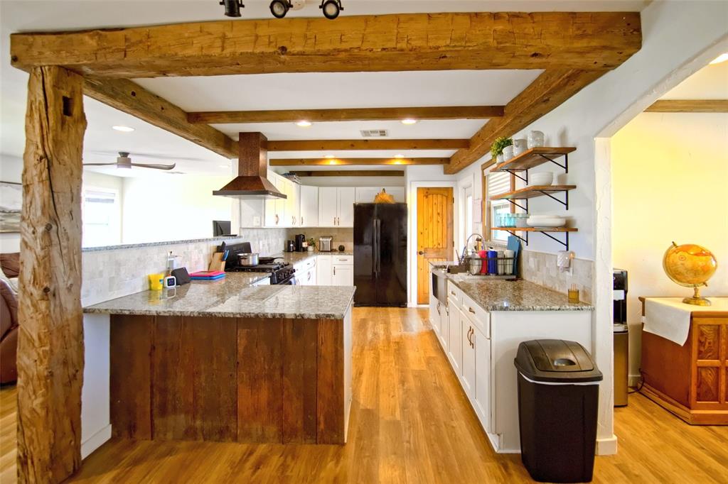 Exposed heavy timber beam ceiling accents, kitchen island with wood-paneled base, stone-style countertops, recessed ceiling lighting, and open-shelf wall storage