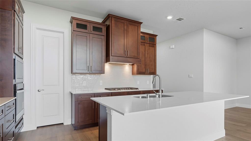 Kitchen featuring stained wood cabinetry, quartz countertops, a central island with an integrated sink, a gas cooktop, and built-in double wall ovens