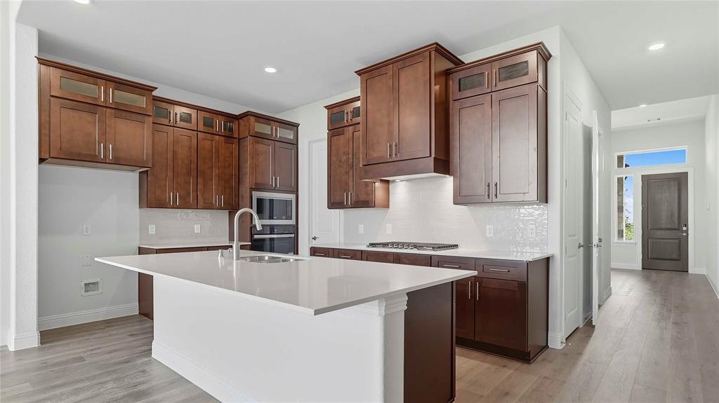 Kitchen featuring cabinetry with glass-paneled upper doors, expansive quartz countertops, an integrated cooktop, built-in wall oven, and wood-finish flooring
