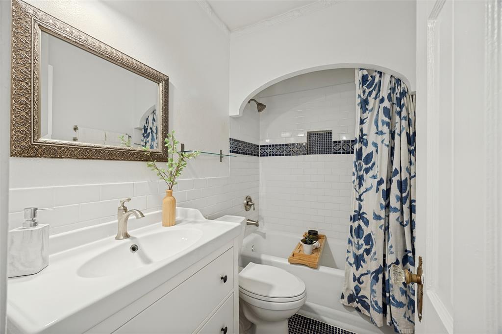 White vanity featuring an integrated sink with a metallic faucet, wall-mounted mirror with a textured metallic frame, tub-shower combination with white subway tile surround, decorative accent tile border, and an arched architectural opening