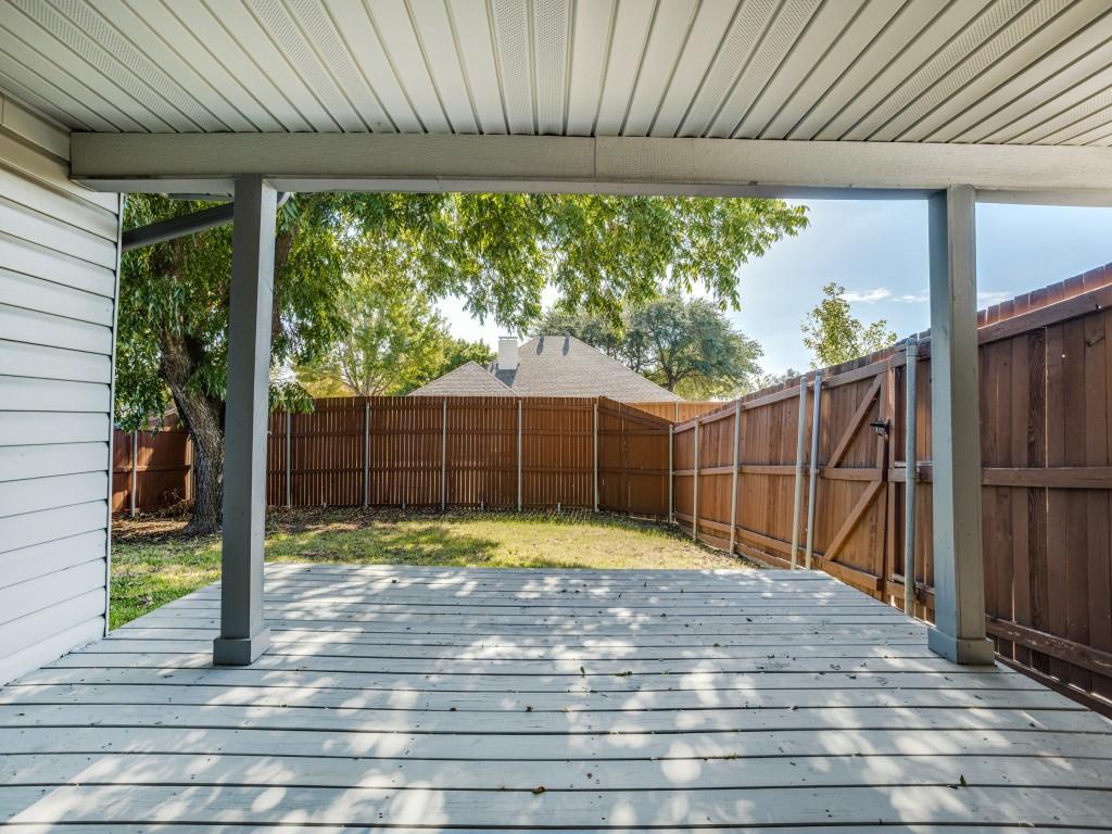 Covered patio with wood-finish decking and structural pillars, encompassing a fenced backyard featuring a grassy lawn and mature trees