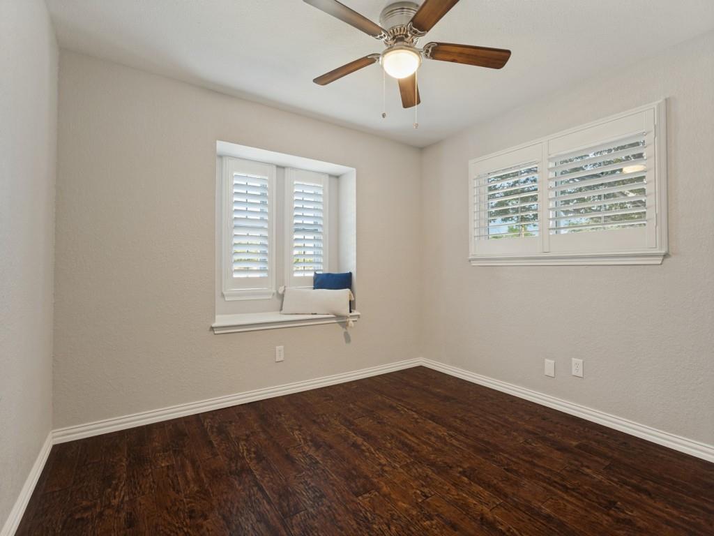 Neutral-toned room featuring dark wood-finish flooring, a ceiling fan with integrated lighting, plantation shutters, and a built-in window seat