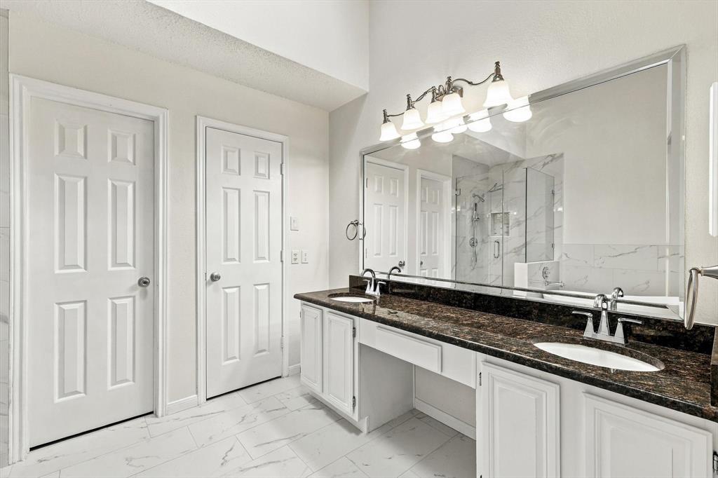 Dual-sink vanity with dark stone countertop, white shaker-style cabinetry, large beveled-edge mirror, and five-light vanity fixture