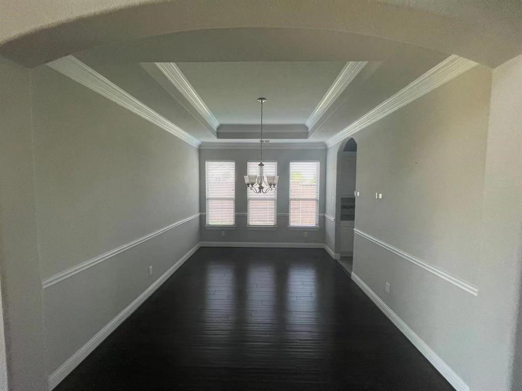 Interior space featuring dark wood-finish flooring, a tray ceiling with crown molding, chair rail trim, and three windows with neutral blinds beneath a central pendant light fixture
