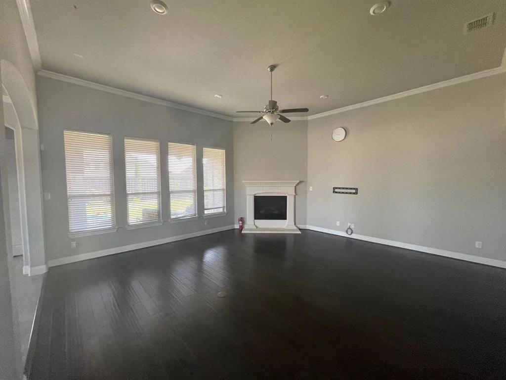 Living area featuring wood-finish flooring, a corner fireplace with a decorative mantel, crown molding, a central ceiling fan, and four oversized windows with horizontal blinds