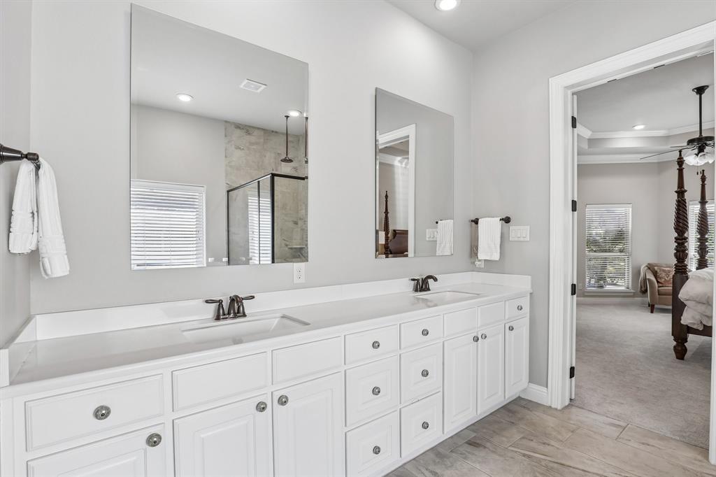 Primary bath featuring dual-sink vanity with white cabinetry, light-toned stone-style countertop, rectangular undermount sinks, and dark metal hardware paired with wood-finish tile flooring