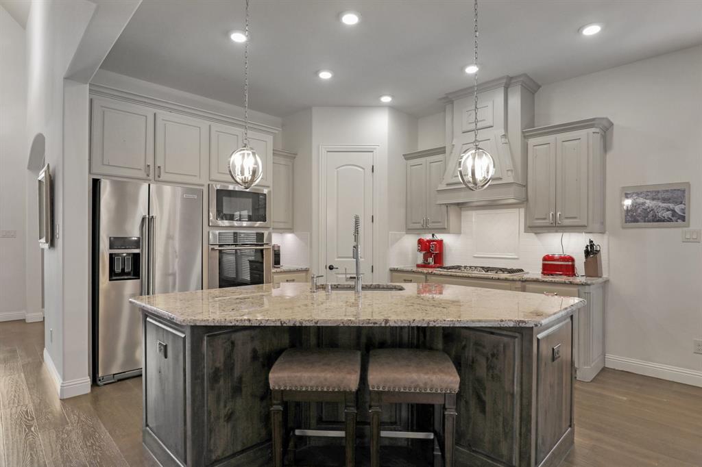 Chef's kitchen featuring light-toned cabinetry, stone-surfaced center island, integrated stainless steel appliances, decorative pendant lighting, and wood-finish flooring Chef's kitchen featuring light-toned cabinetry, stone-surfaced center island, integrated stainless steel appliances, decorative pendant lighting, and wood-finish flooring
