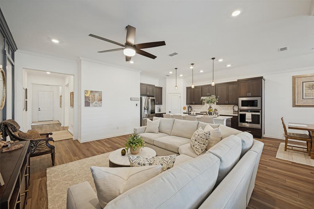Open-concept interior featuring wood-finish flooring, recessed ceiling lighting, a contemporary multi-blade ceiling fan, and a kitchen equipped with dark cabinetry, stone countertops, and built-in stainless steel appliances Open-concept interior featuring wood-finish flooring, recessed ceiling lighting, a contemporary multi-blade ceiling fan, and a kitchen equipped with dark cabinetry, stone countertops, and built-in stainless steel appliances