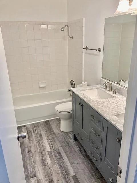 Grey shaker-style vanity with stone-finish countertop, dual-basin under-mount sinks, wood-finish flooring, and white tiled tub surround