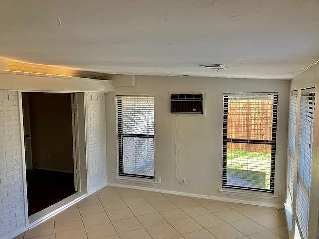 Interior room featuring tile flooring, painted brick wall accents, multiple windows with blinds, and a built-in window air conditioning unit Interior room featuring tile flooring, painted brick wall accents, multiple windows with blinds, and a built-in window air conditioning unit
