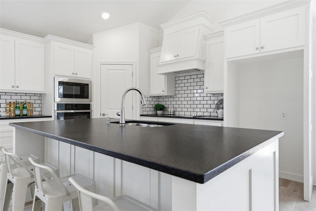 Kitchen featuring a central island with dark solid surface countertops, white cabinetry with crown molding, subway tile backsplash, and a built-in microwave and wall oven Kitchen featuring a central island with dark solid surface countertops, white cabinetry with crown molding, subway tile backsplash, and a built-in microwave and wall oven