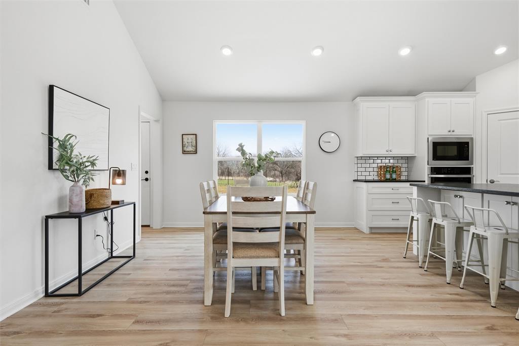 Kitchen and dining area featuring white shaker-style cabinetry, dark countertops, subway tile backsplash, built-in stainless steel double ovens, and wood-finish flooring