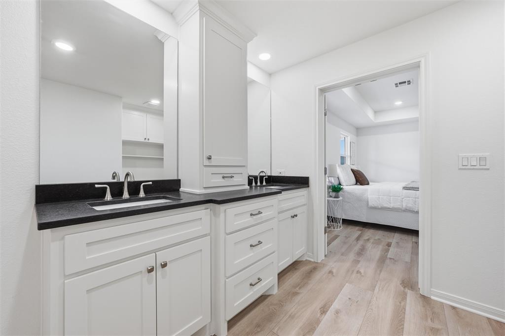 Dual-sink vanity featuring white shaker-style cabinetry, dark solid-surface countertops, integrated medicine cabinetry, and brushed nickel fixtures, complemented by wood-finish flooring