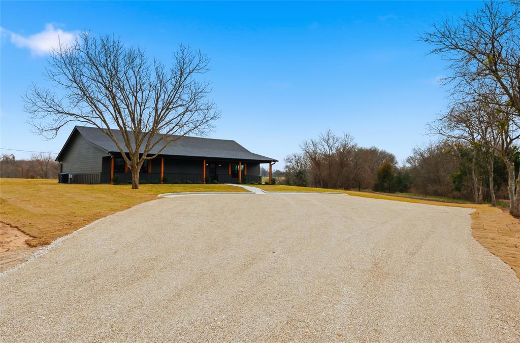 Single-story exterior featuring dark siding, a covered front porch with wooden support posts, a concrete walkway, and a gravel driveway situated on expansive acreage Single-story exterior featuring dark siding, a covered front porch with wooden support posts, a concrete walkway, and a gravel driveway situated on expansive acreage