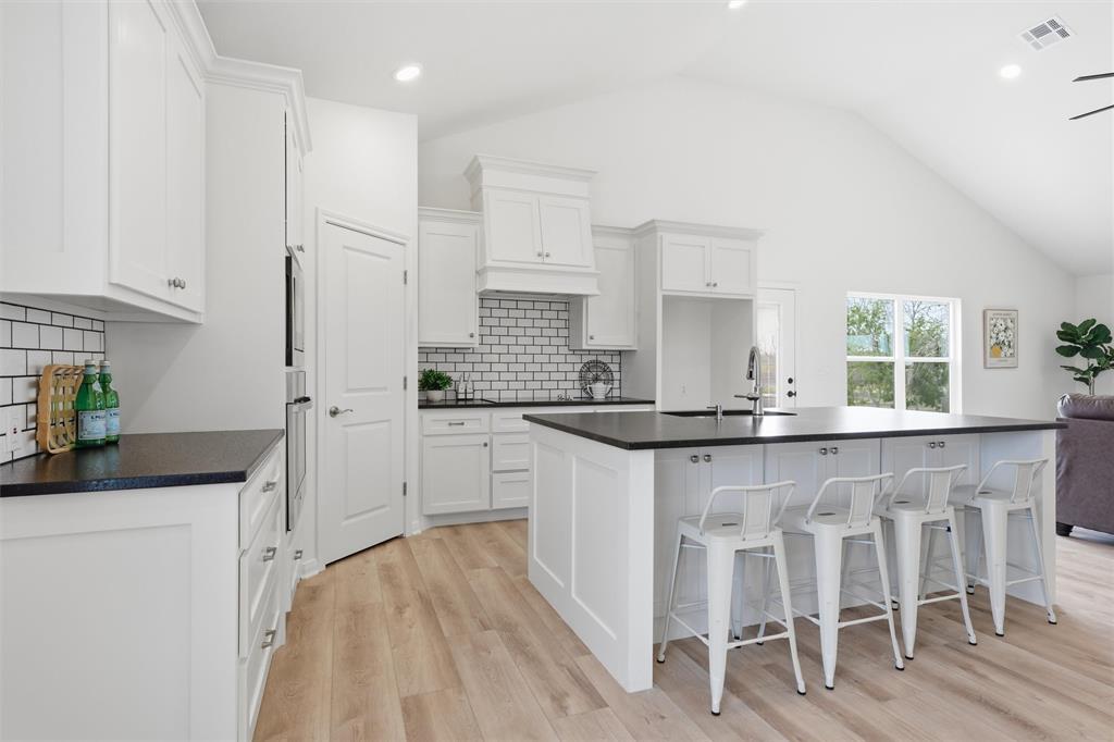 Kitchen featuring vaulted ceilings, white shaker-style cabinetry, dark solid-surface countertops, subway tile backsplash, and wood-finish flooring