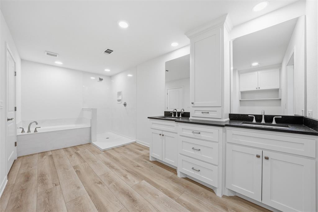 Dual vanity with white cabinetry and dark countertops, wood-finish flooring, built-in storage tower, soaking tub, and walk-in shower