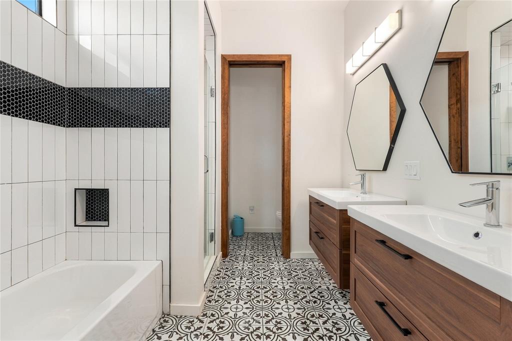 Dual floating vanities with wood-finish cabinetry, patterned tile flooring, hexagonal wall mirrors, and a tiled shower surround with a built-in recessed niche