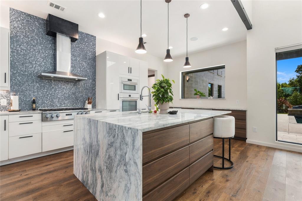 Kitchen featuring a central island with a stone waterfall countertop, wood-finish cabinetry, built-in double wall ovens, stainless steel range hood, and textured mosaic tile backsplash