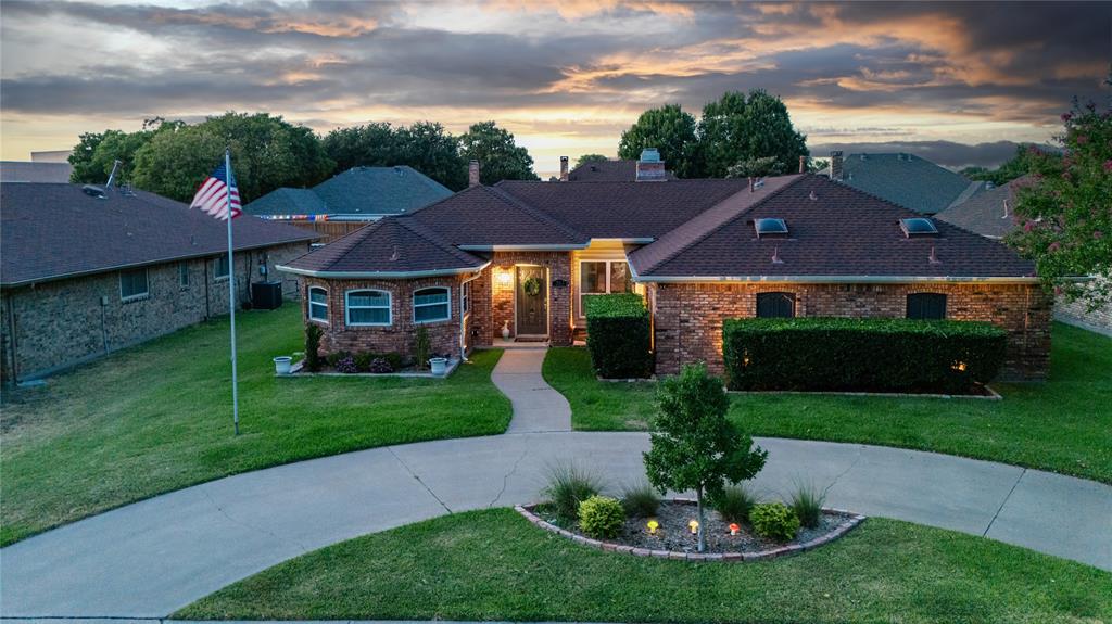 Brick Colonial-style residence featuring a welcoming front entrance with illuminated architectural lighting, manicured lawn with mature landscaping, circular concrete driveway, and an American flag display