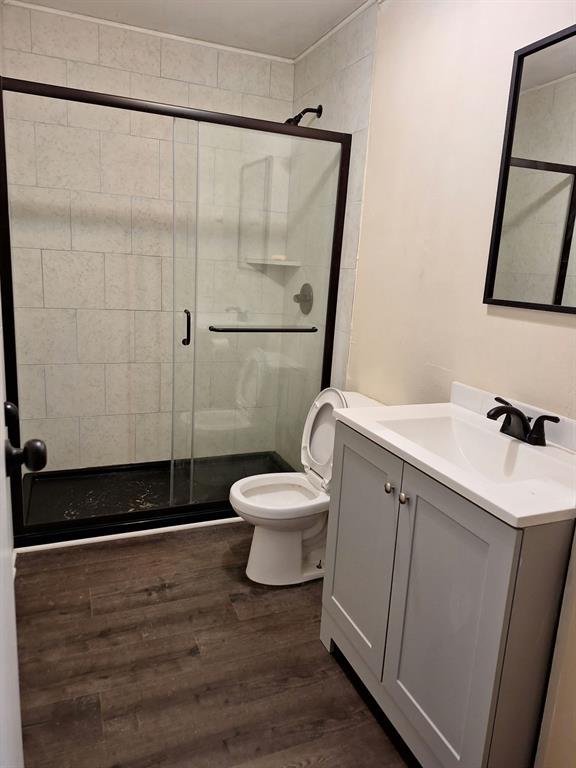 Bathroom featuring a walk-in shower with a sliding glass door, a dark-toned shower base, light-colored tile wall surround, wood-finish flooring, and a gray vanity with a white countertop