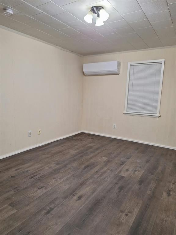 Interior room featuring wood-finish flooring, a wall-mounted ductless heating and cooling unit, a single window with blinds, and a multi-light flush-mount ceiling fixture